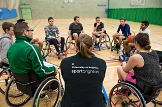 Wheelchair sports players in a circle inside a gym. The back of one person's shirt has the Sport Brighton University of Brighton logo