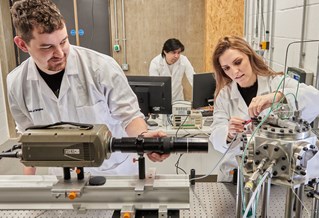 Three researchers wearing white lab coats, working with machinery and digital screens in the University of Brighton Advanced Engineering Centre
