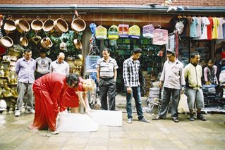 Two performance artists, dressed in bright red saris, push two large blocks of ice along a street in Nepal. In the background Nepali shop with wares and onlookers.