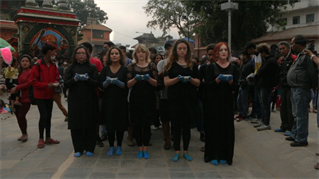 Urban square in Nepal. Five artists walking in a side-by-side row, dressed in black. They each carry a small bowl of milk in raised cupped, blue-gloved hands. Looking straight ahead. Spectators to the left and right and behind.