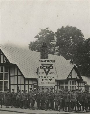 Black and white photograph. Military men in World War One uniforms stand on the pavement outside a single-storey mock-Tudor building. A large sign reads: Shakespeare YMCA rest and recreation hut for men of HM Forces. Popularly known as the Shakespeare Hut