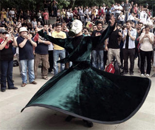 A crowd in contemporary park setting watch a performer with a wide spread skirt in green velvet. A Whirling Dervish.