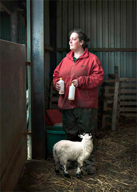 Colour photograph by Zoe Childerley. Female farmer stands in a barn with a lamb at her feet, holding two bottles of milk. Photographs from The Debatable Lands publication