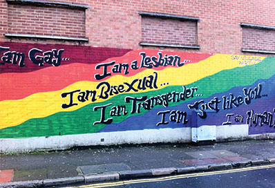 A rainbow painted wall with 'I am Gay/I am Bisexual/I am Transgender/I am just like you' graffitied on it.