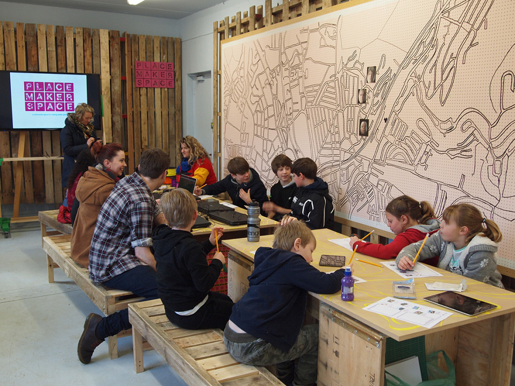 Group of school children sat at a wooden bench, drawing and discussing with large map on wall behind.