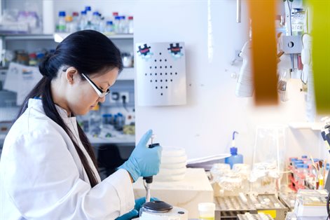 A student wearing a lab coat and gloves using a pipette at a laboratory workstation with equipment and supplies visible in the background.