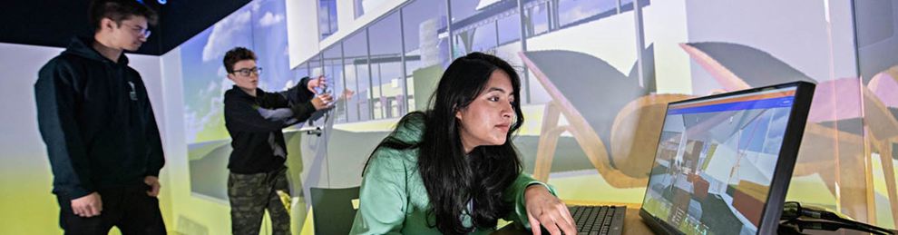 Female student working a computer with two students in the background discussing visual of a building on a large screen