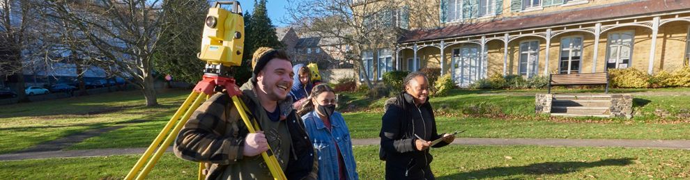 Three students walking outside carrying a total station