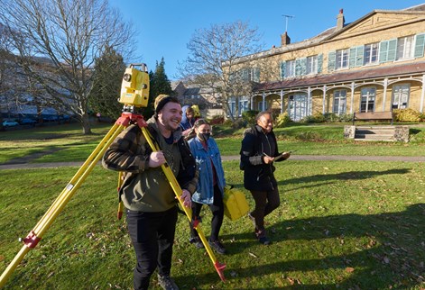 Three students walking outside carrying a total station