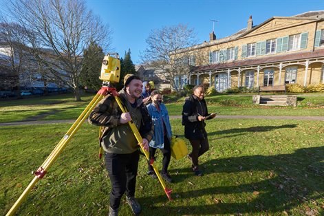 Three students walking outside carrying a total station