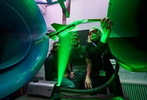 Lecturer demonstrating wind tunnel to two students wearing goggles with laser green light