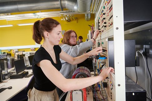 Two students standing up using large vertical electrical engineering equipment with cables