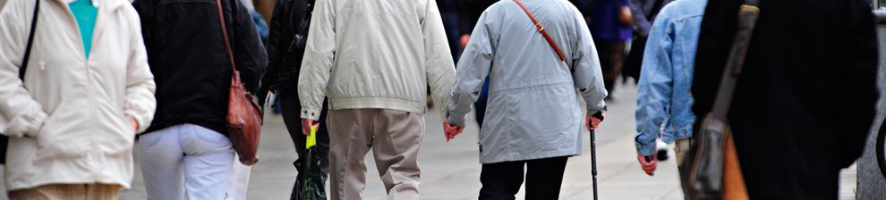 Banner representing the study of Sociology for PhD, shows a low level camera view of people walking in a town. An elderly couple hold hands, others walk past with hands in pockets.