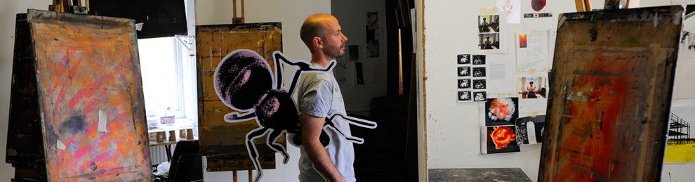 A student standing between two canvases with a printed cut out of a giant ant under his arm