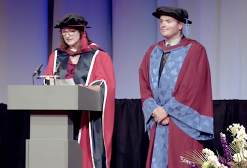 Graduation ceremony, two figures in red and blue robes on stage at the University of Brighton