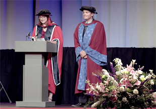 Graduation ceremony, two figures in red and blue robes on stage at the University of Brighton