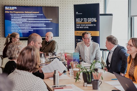 Help To Grow classroom with group of people in discussion around a table and a Help To Grow banner stand