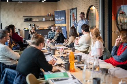 Help To Grow classroom with people sat at a long table and a speaker talking to the group