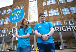 Two open day helpers in blue T-shirts holding a sign that reads 'Here to help'