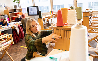 Student in a workroom reaching for spools of colourful thread.