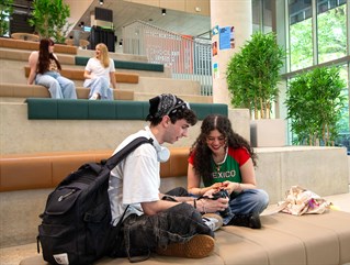 Two students looking at a camera together on the stepped seating in the Elm House foyer