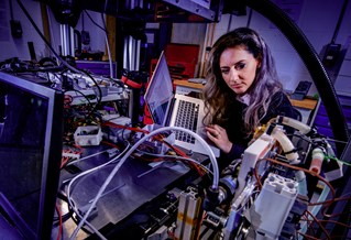 Female engineering researcher in laboratory. She holds a laptop while examining complex mechanical surroundings under pale violet light.