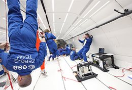 A scene from a parabolic flight showing crew in blue boiler-suits marked ESA zero G team, floating in a long white tubular space, some upside down. Parabolic micro-gravity images provided by European Space Agency.