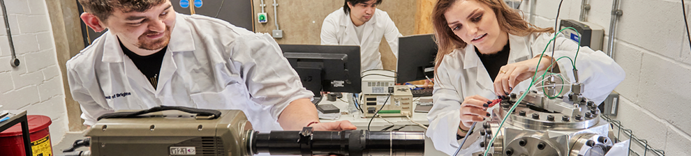 Three researchers in white lab-coats work with complex machinery and dual computer screens inside the Advanced Engineering Centre building.