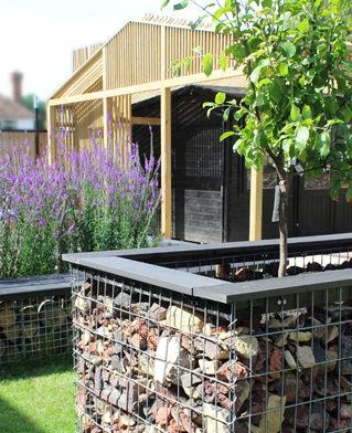 Wooden open shelter in a community garden with barriers built of stone and log fillings of mesh.