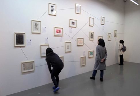 Exhibition wall nineteen small framed images randomly arranged with drawn lines between them. Three gallery visitors examining.