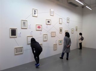 Exhibition wall nineteen small framed images randomly arranged with drawn lines between them. Three gallery visitors examining.