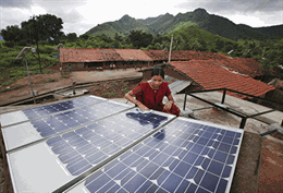 Woman in sari sits alongside a large solar panel with bungalows behind her and mountains in the distance.