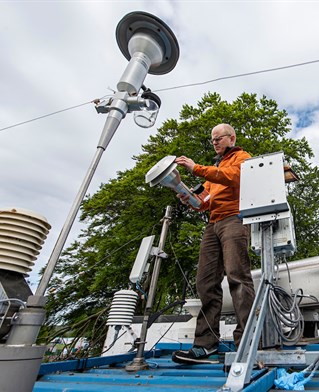 The Brighton Atmospheric Observatory air monitoring station. Earth observation scientist in high vis jacket stands on an urban rooftop with leafy backdrop. The chrome equipment surrounding him includes large tubes and tall antennae.