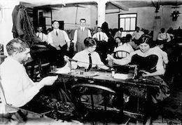 Black and white indoor image of working group. Long tables. Two women at a table with threads and equipment dominate. Further tables and men in waistcoats and ties.