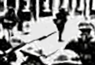Very grainy de-focused black and white image showing soldiers in a wide terraced street. Middle ground standing with rifles, foreground walking towards camera with helmets and rifles. Civilians on pavement and walking across the road. Image of Belfast str