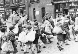 Black and white photograph of a  street scene with children in shorts and cloth caps crossing in a queue, each carrying large, informally made backpacks