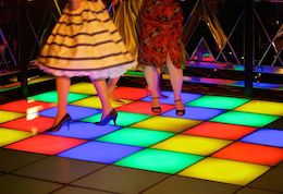Colour photograph of dancers, showing two figures in vintage dresses and shoes on a lit panel colour-block dancefloor.