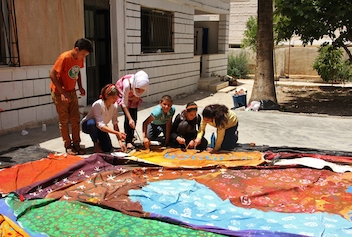 Grainy coloured photograph. Children together with older figures examine brightly coloured cloth outdoors.