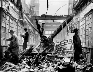 Black and white press photograph of three men inspecting the intact bookshelves of the bombed library while the floor is covered in debris and the remains of the fallen ceiling, now open to the air. Holland House library after an air raid