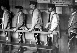 Four men stand, each wearing formal hats and shirts. They each show one bare leg and the closer leg shows a strapping or mechanism down it.