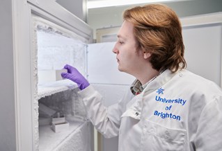 Scientist with white coat and purple gloves working in genomics lab with large refrigeration unit