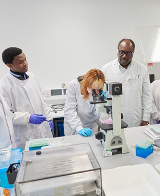 University of Brighton Genomics lab. Scientists in white coats group around a microscope.