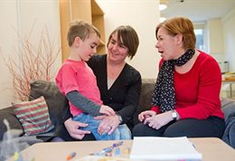 A small boy sitting on a woman's lap while another woman chats to him depicting resilience research and practice.