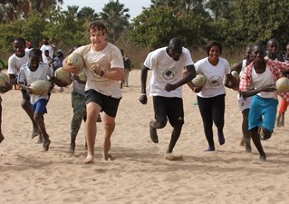 Group of boys and men running towards the camera across a beach, one with a rugby ball. Rugby 4 Peace.
