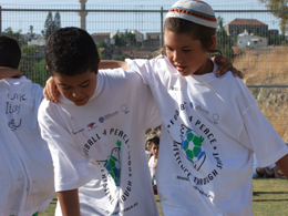 Two boys link arms, one with a kippah on his head, wearing Football4peace white teeshirts. Hot weather and sharp shadow with sun in background, pointing to a football at their feet.