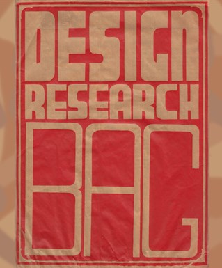 A printed paper bag with pale cream capital lettering on red background, reads Design Research Bag