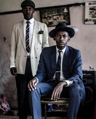 Grainy photograph of two black male models in fashionable suits and hats. Indoors, bare floorboards and pale pink walls. Seated man in blue, standing man wears a white jacket.