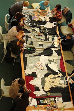 High viewpoint of a group seated around a long table each sewing a section of a tapestry replica of Picasso's Guernica