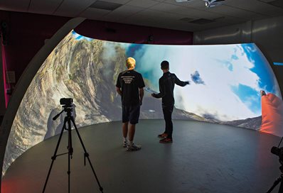 Two researchers stand within a quarter sphere on which is a 3D projection of a scene with mountains, sky and clouds.