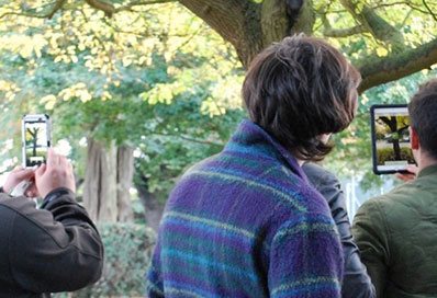 Digital researchers collaborate in an outdoor, wooded environment using integrated phone apps yielding digital information on the environment.